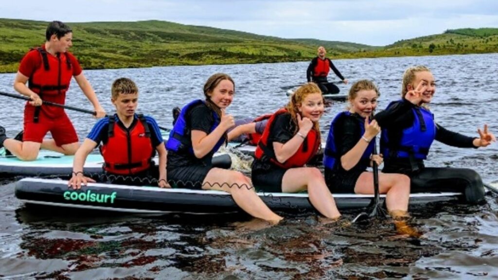 7 teenagers in life vests and wet suit are in a paddle boat smiling and having a great time