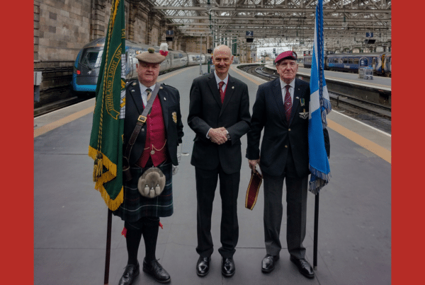 two military men with flags and Vice Dean of The Merchants House Andrew MacFarlane stand on a platform in Central Station, Glasgow