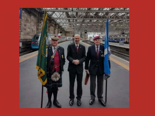 two military men with flags stand with Andrew on the platform at Glasgow Central Station