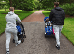 two young men push prams through a park with their back to the camera