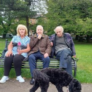 a park bench with three older adults having a chat whilst a dog wanders at their feet