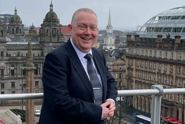 CEO Craig Vickery is photographed on the rooftop of The Merchants House with a view of George Square behind him. He wears a dark suit with a blue shirt.