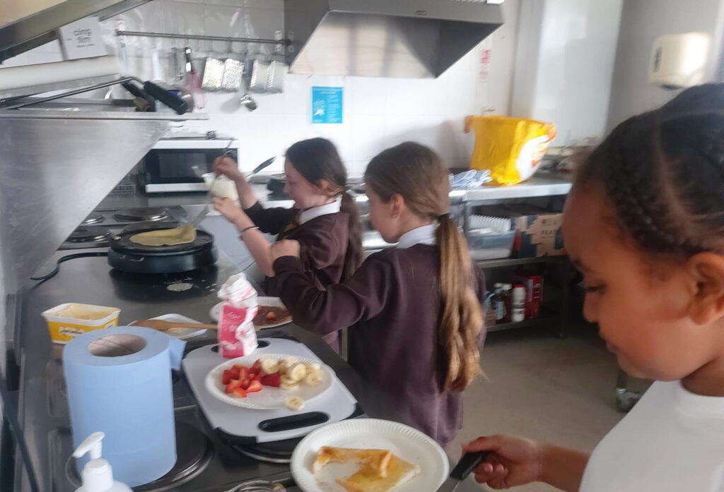 three young girls make pancakes in the cafe kitchen with a plate of chopped bananas and strawberries as a topping