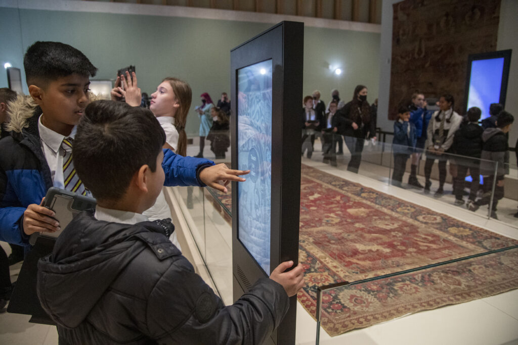 Children touching an interactive screen at The Burrell Collection in front of a huge historic rug