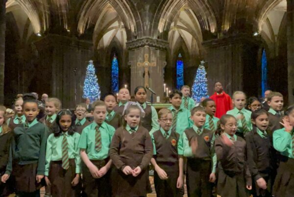 A photo of a children's choir standing in a historic building with pointed arches behind them. The children are singing and are wearing school uniform. The lighting is low.