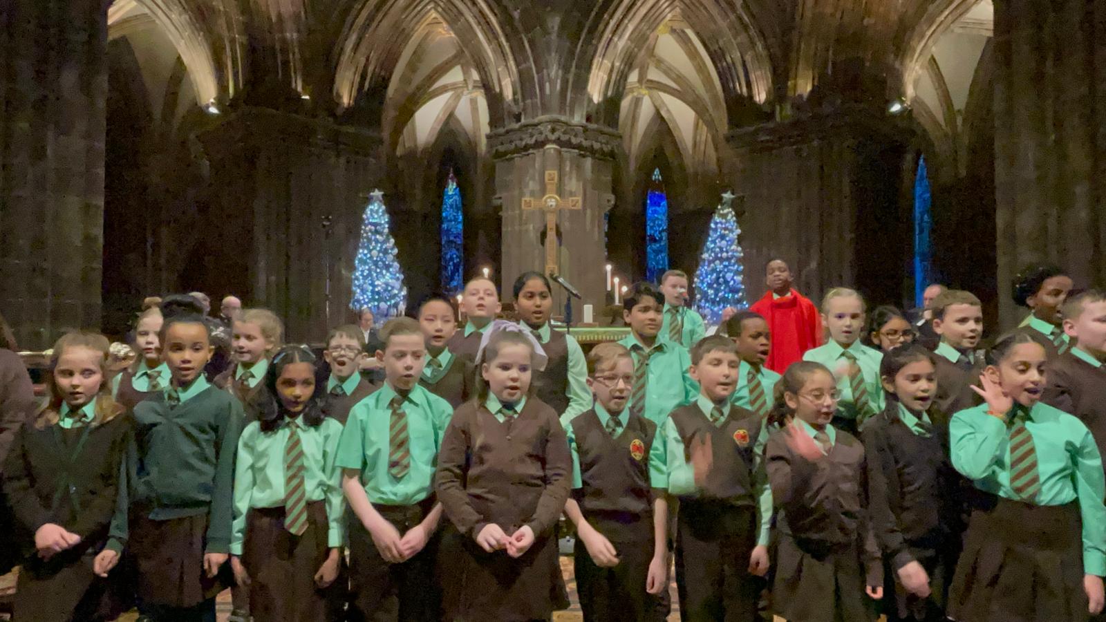 A photo of a children's choir standing in a historic building with pointed arches behind them. The children are singing and are wearing school uniform. The lighting is low.