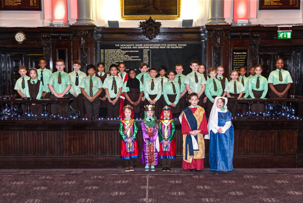 A choir of young children in school uniform set against a backdrop of the interior of a historic building.