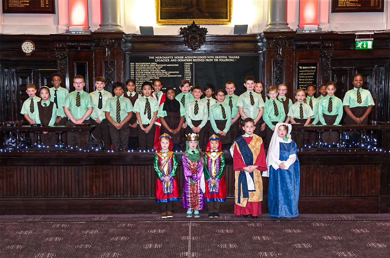 A choir of young children in school uniform set against a backdrop of the interior of a historic building.